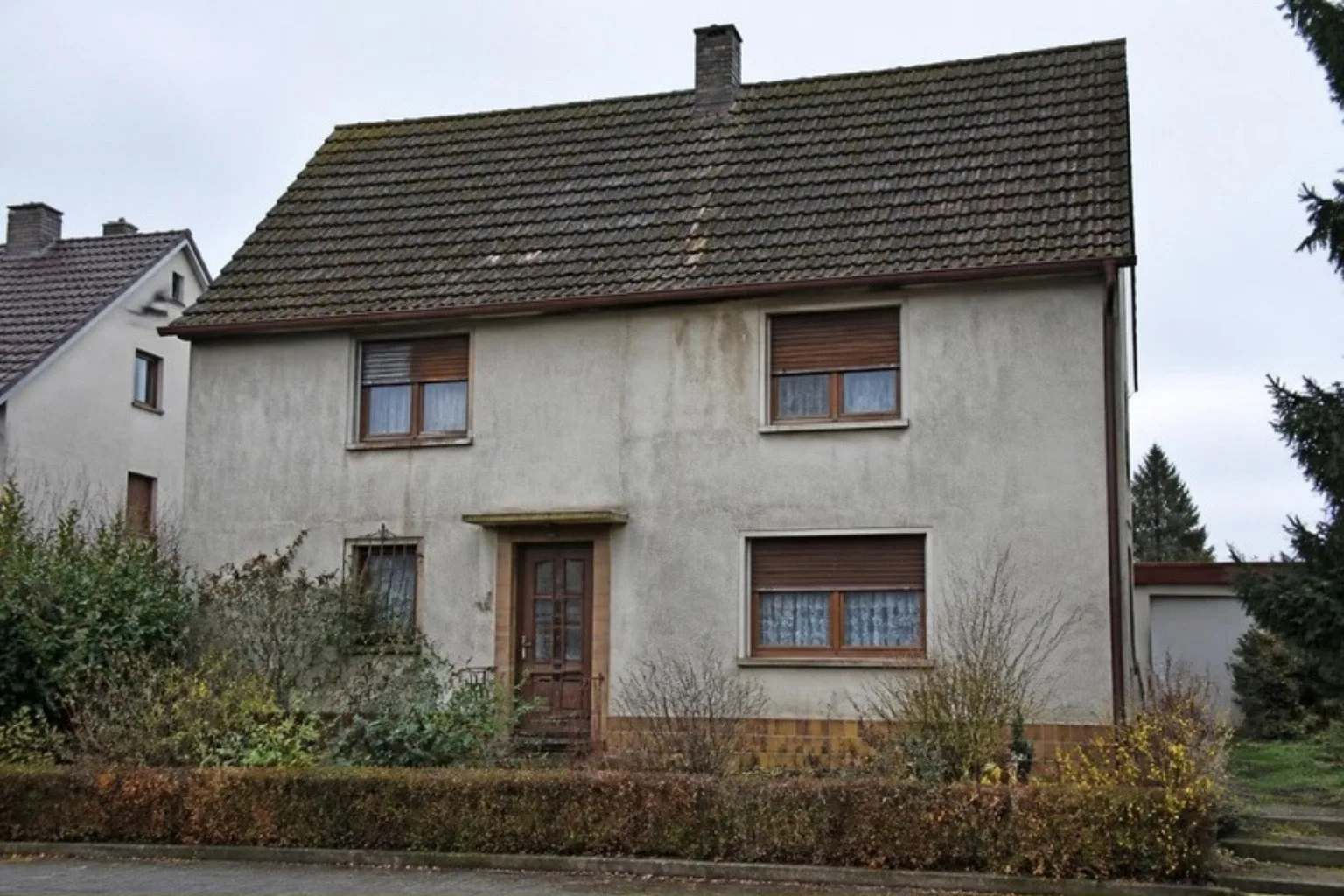Einfamilienhaus aus den 1960er Jahren vor der Sanierung: graue rissige Fassade, alte Holzfenster, bemoostes Dach
